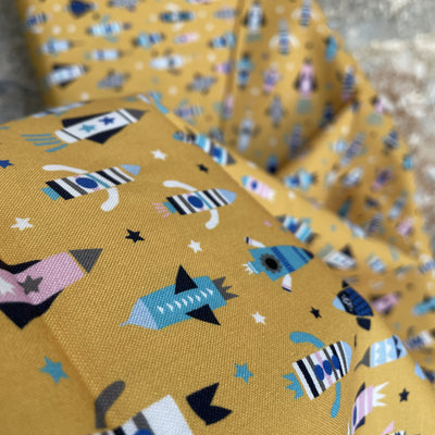 A mustard base fabric with rockets and stars in pink, white, black, blue and turquoise. Blast off fabric by Dashwood Studios 